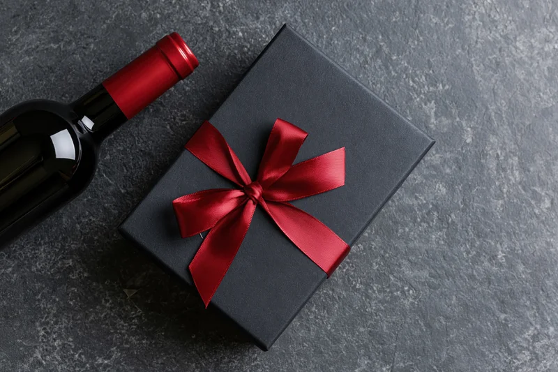 Dark gift box with red satin ribbon bow beside a wine bottle on stone surface