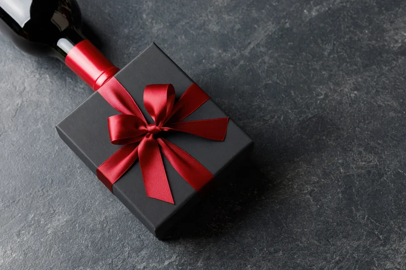 Dark gift box with red satin ribbon bow beside a wine bottle on slate surface