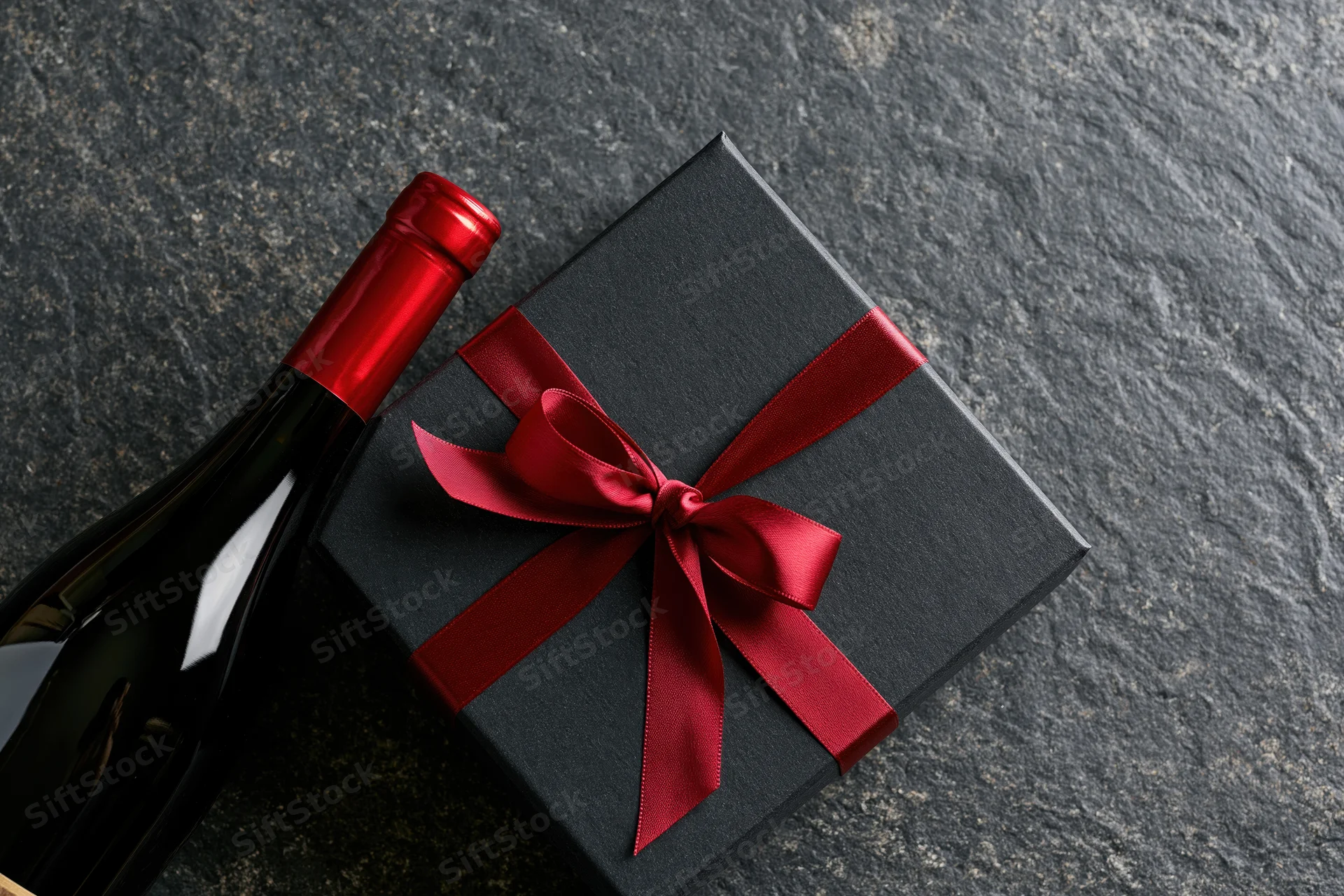 Dark gift box with red satin ribbon bow beside a wine bottle on stone surface