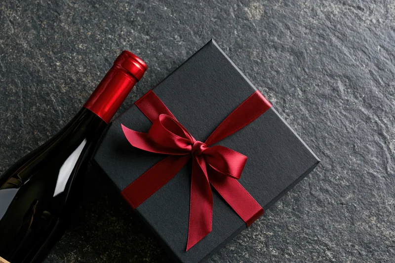 Dark gift box with red satin ribbon bow beside a wine bottle on stone surface
