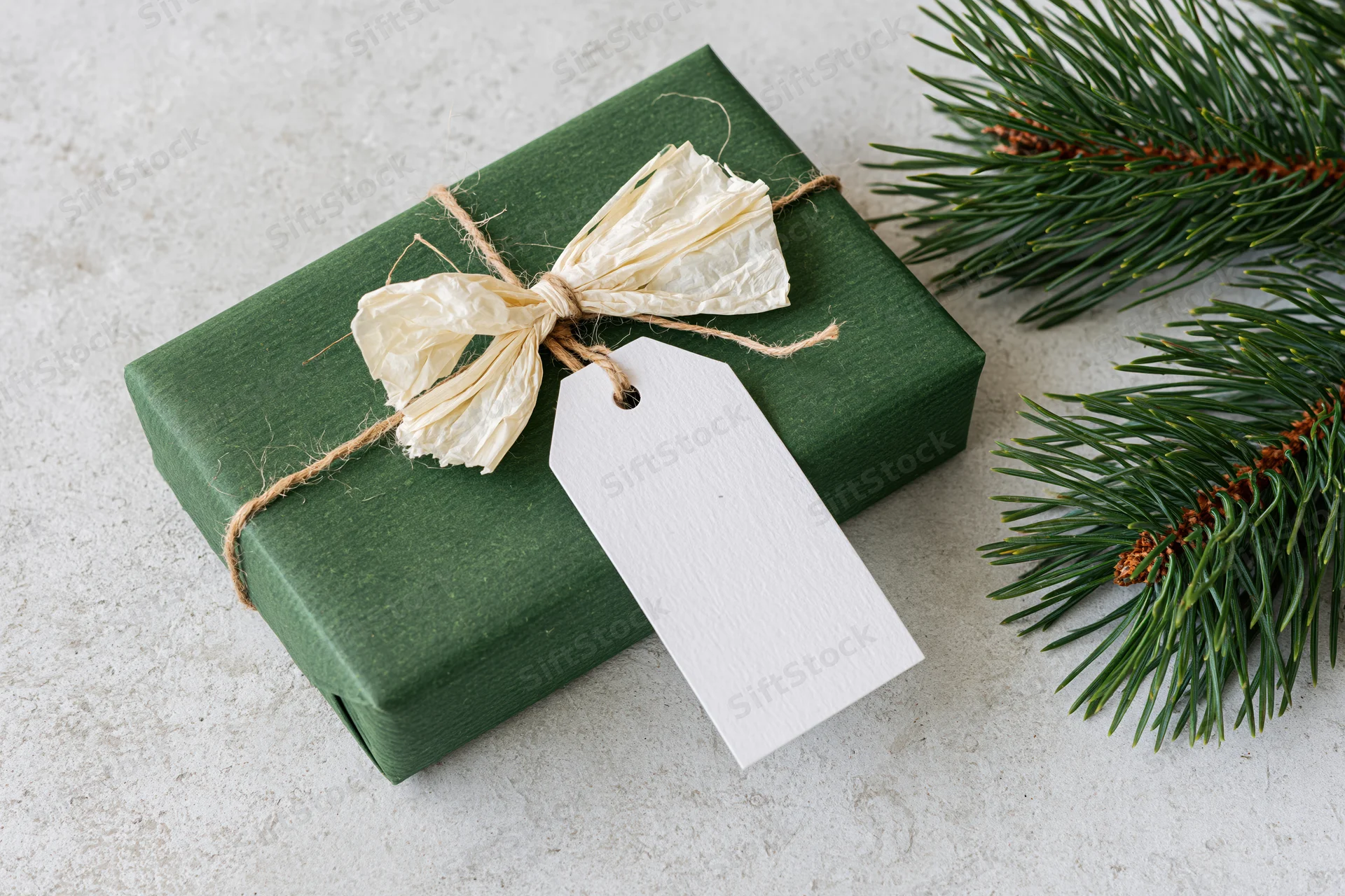 Dark green gift box wrapped in kraft paper with raffia bow and white tag beside pine branches on light surface
