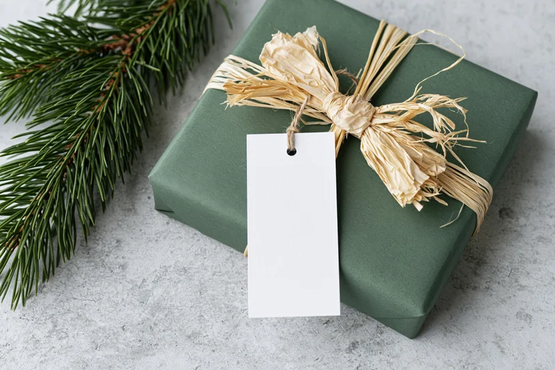 Dark green gift box wrapped with raffia bow and blank tag beside pine sprig on gray surface