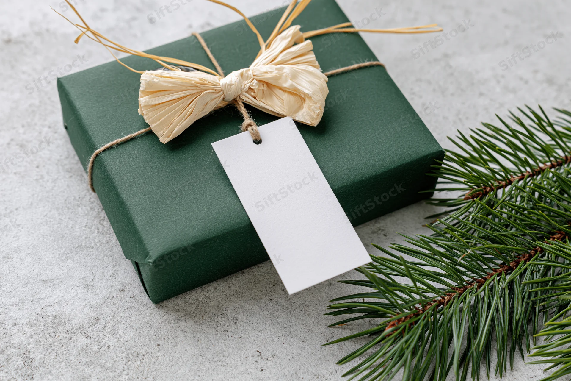 Dark green gift box wrapped with raffia bow and blank tag beside pine branch on textured surface