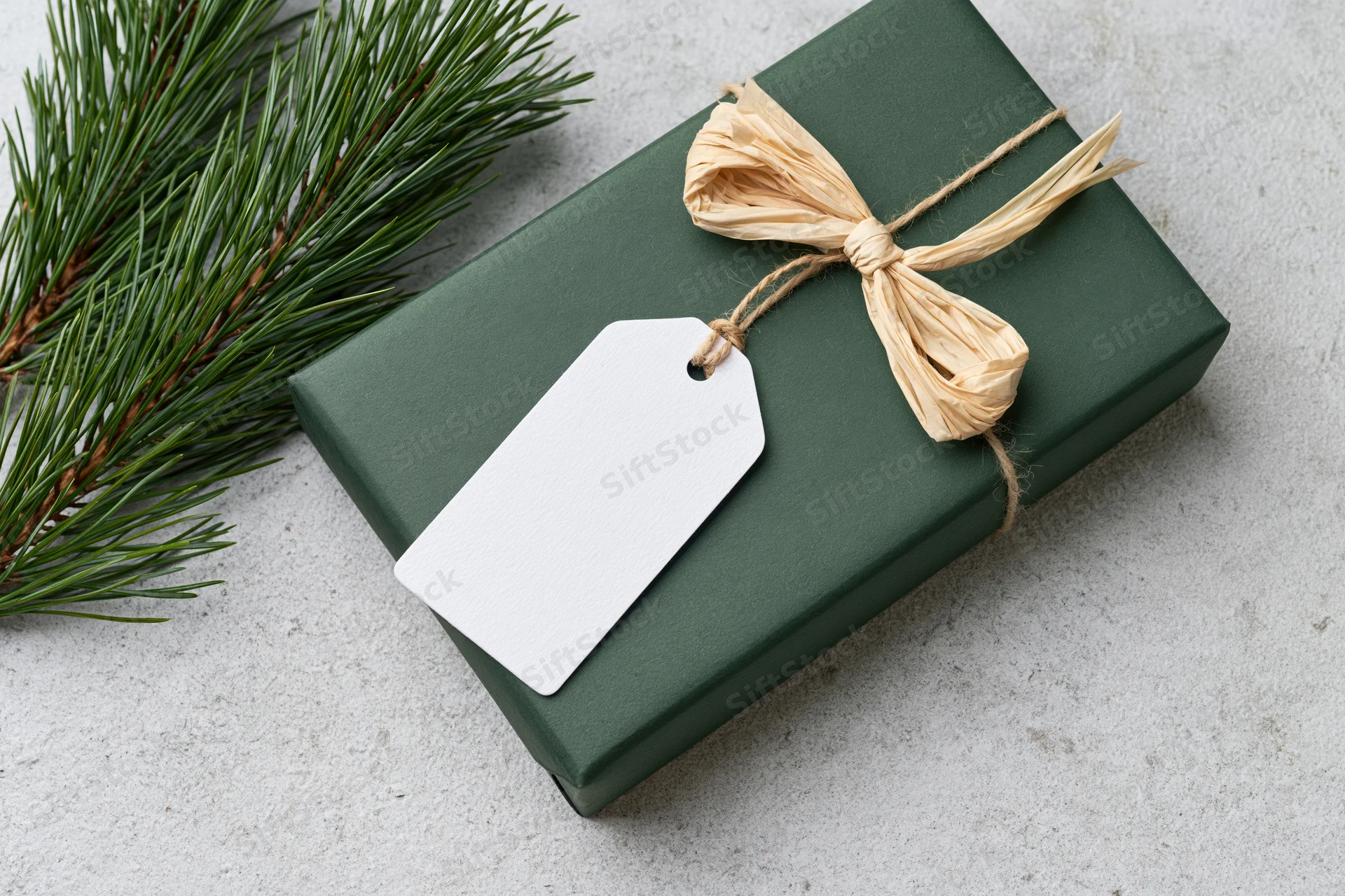 Dark green gift box wrapped with raffia bow and blank tag beside pine branch on concrete surface