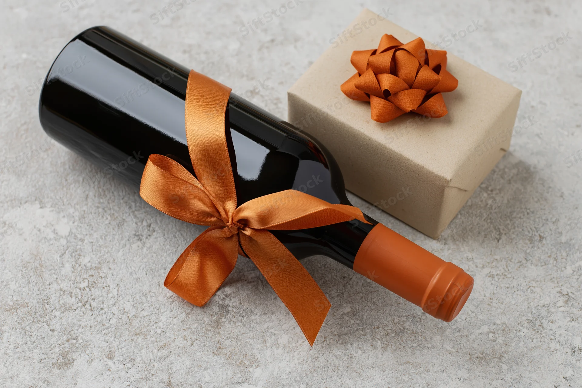 Dark wine bottle with orange satin ribbon lying beside a gift box on textured surface