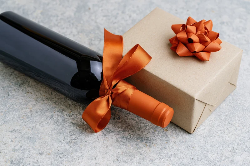 Dark wine bottle with orange satin ribbon beside a kraft paper gift box on stone surface