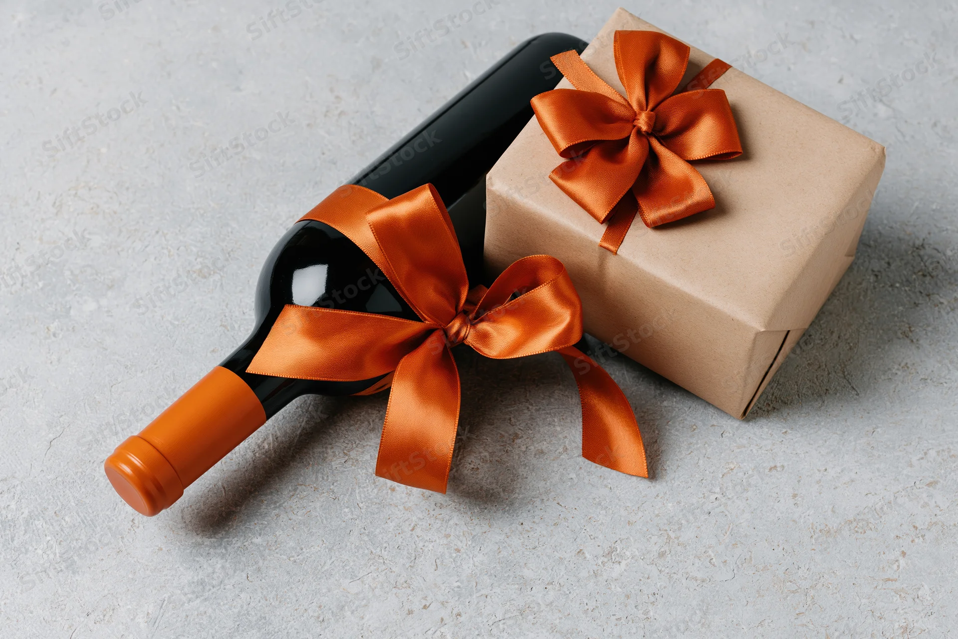 Dark wine bottle and gift box wrapped with orange satin ribbons on gray surface