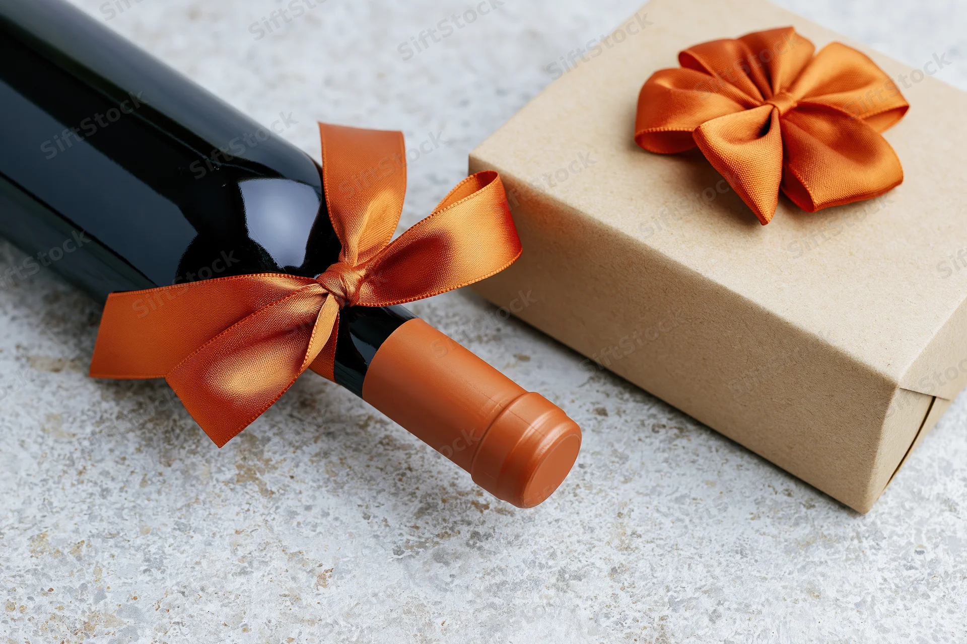 Dark wine bottle with orange satin ribbon beside a kraft paper gift box on textured surface