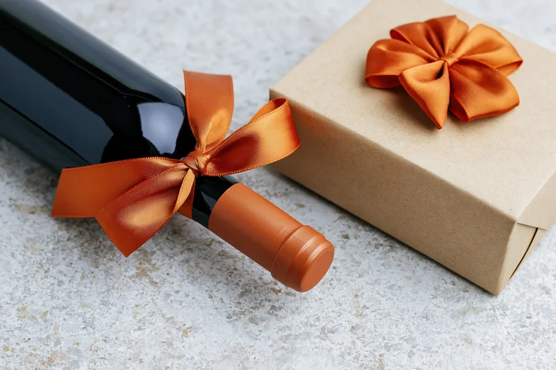 Dark wine bottle with orange satin ribbon beside a kraft paper gift box on textured surface