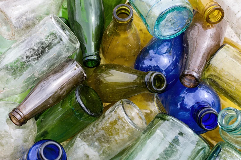 Pile of empty glass bottles in various colors and shapes, some broken and dirty, arranged closely together in a heap