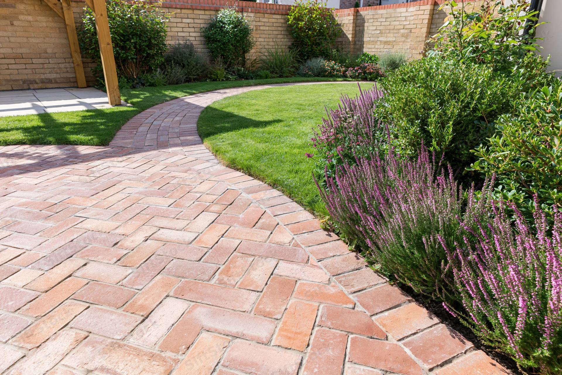 Curved brick pathway winding through a landscaped backyard garden with green lawn and purple flowering shrubs