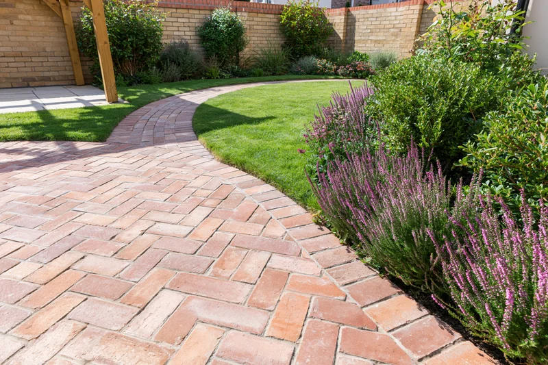Curved brick pathway winding through a landscaped backyard garden with green lawn and purple flowering shrubs