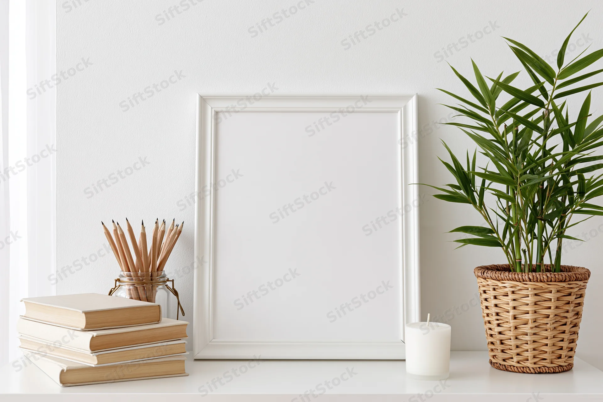 Empty white frame leaning on a shelf beside books, a bamboo plant in a wicker pot, and a candle