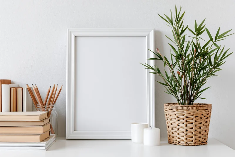 Empty white frame leaning on a white desk beside books, pencils, candles and a bamboo plant in a wicker basket