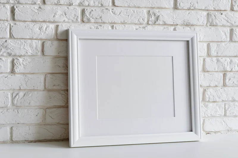 White picture frame with mat insert leaning against white brick wall on shelf