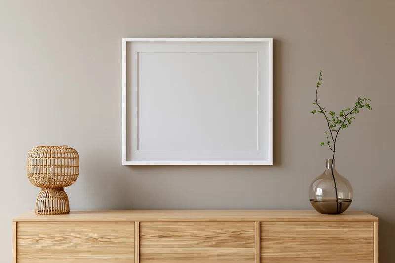Empty white frame hanging on warm greige wall above wooden cabinet with vase and lamp