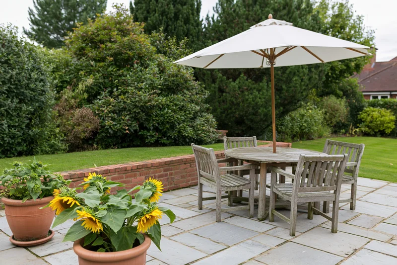 Weathered teak outdoor dining set under umbrella on stone patio with potted sunflowers and green lawn