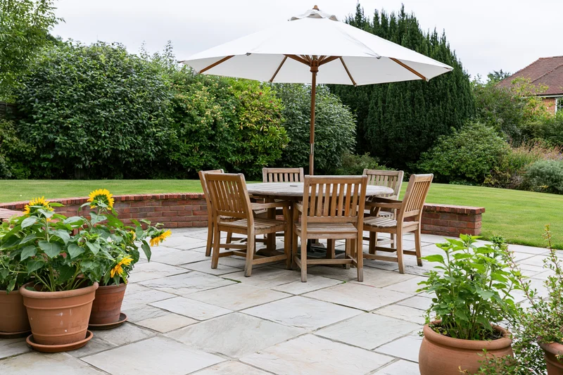 Wooden patio furniture with umbrella and potted sunflowers on stone tiles surrounded by green lawn and hedge