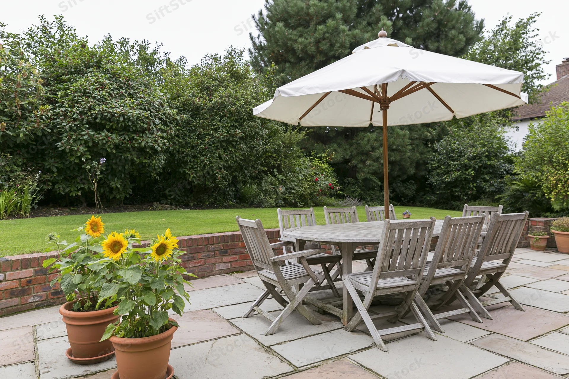 Weathered teak outdoor dining set with umbrella on stone patio surrounded by greenery and potted sunflowers