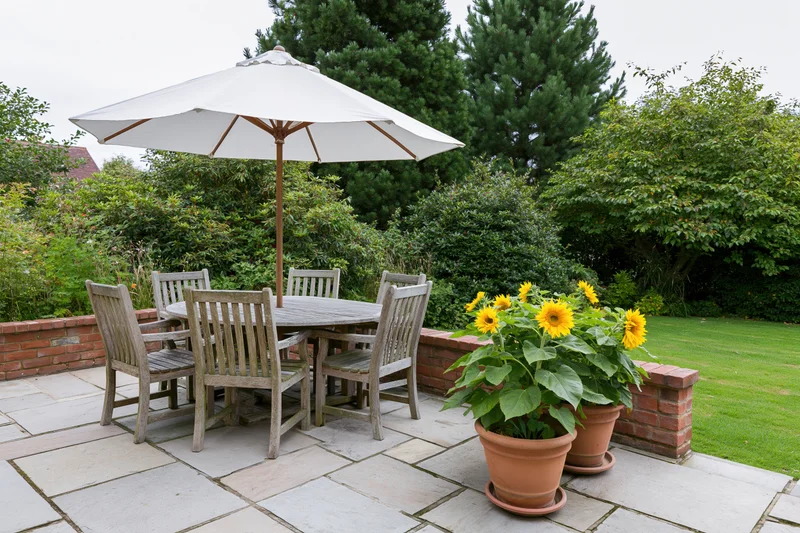 Weathered teak outdoor dining set under umbrella on stone patio with potted sunflowers and greenery