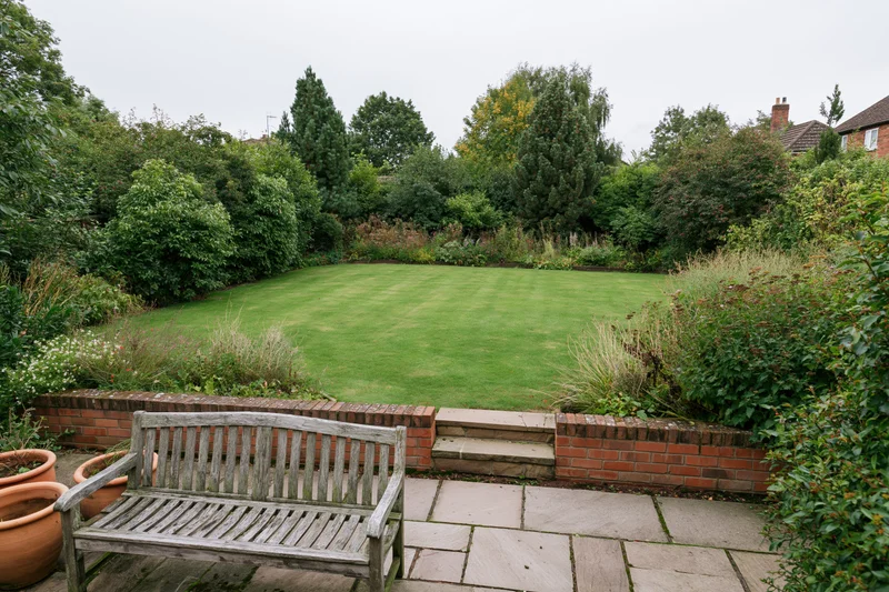 Elevated view of a suburban backyard garden with a wooden bench on a stone patio overlooking a green lawn bordered by shrubs and trees