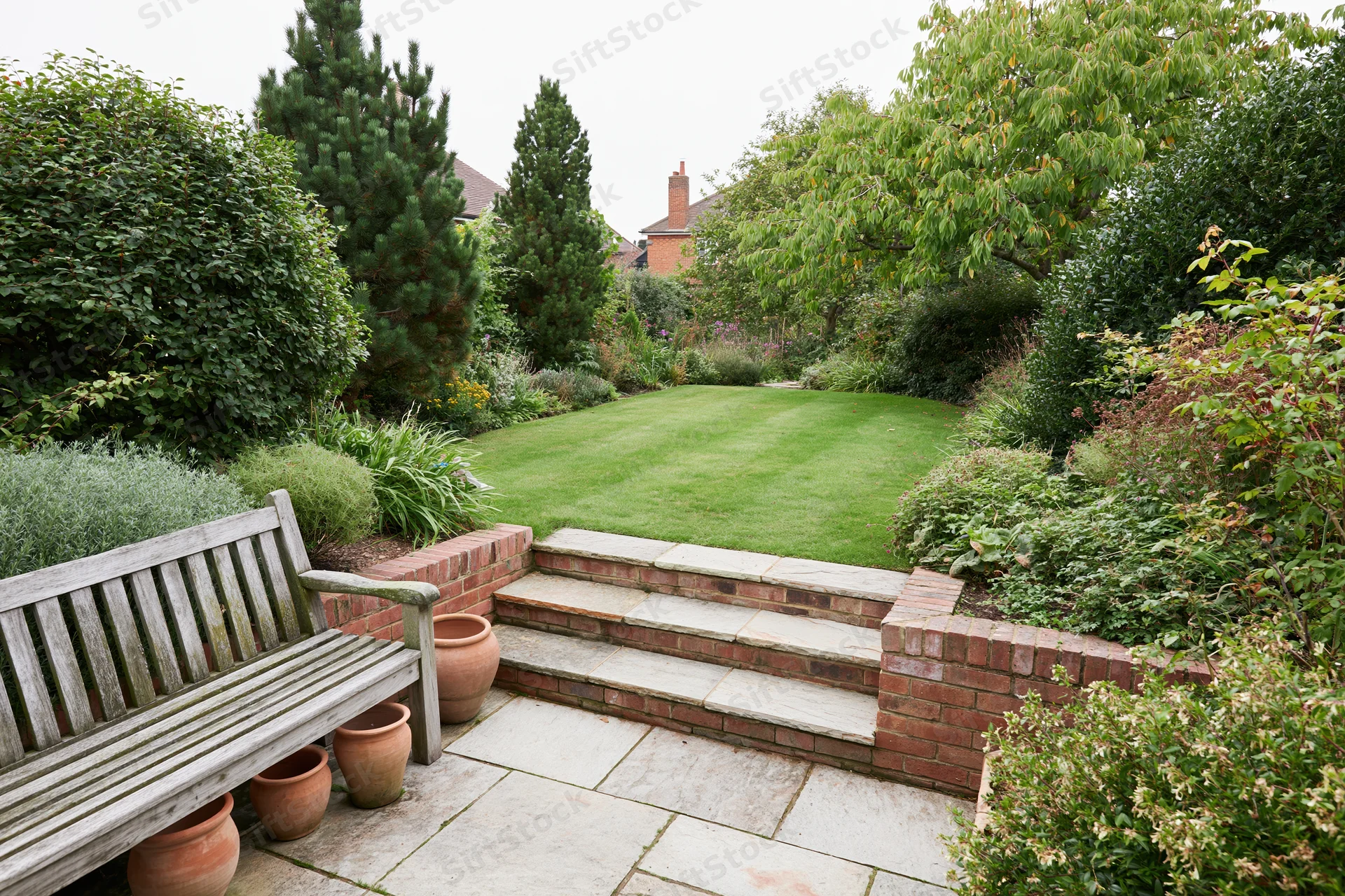 Elevated patio overlooking a landscaped suburban garden with stone steps, brick retaining walls, and a wooden bench beside terracotta pots