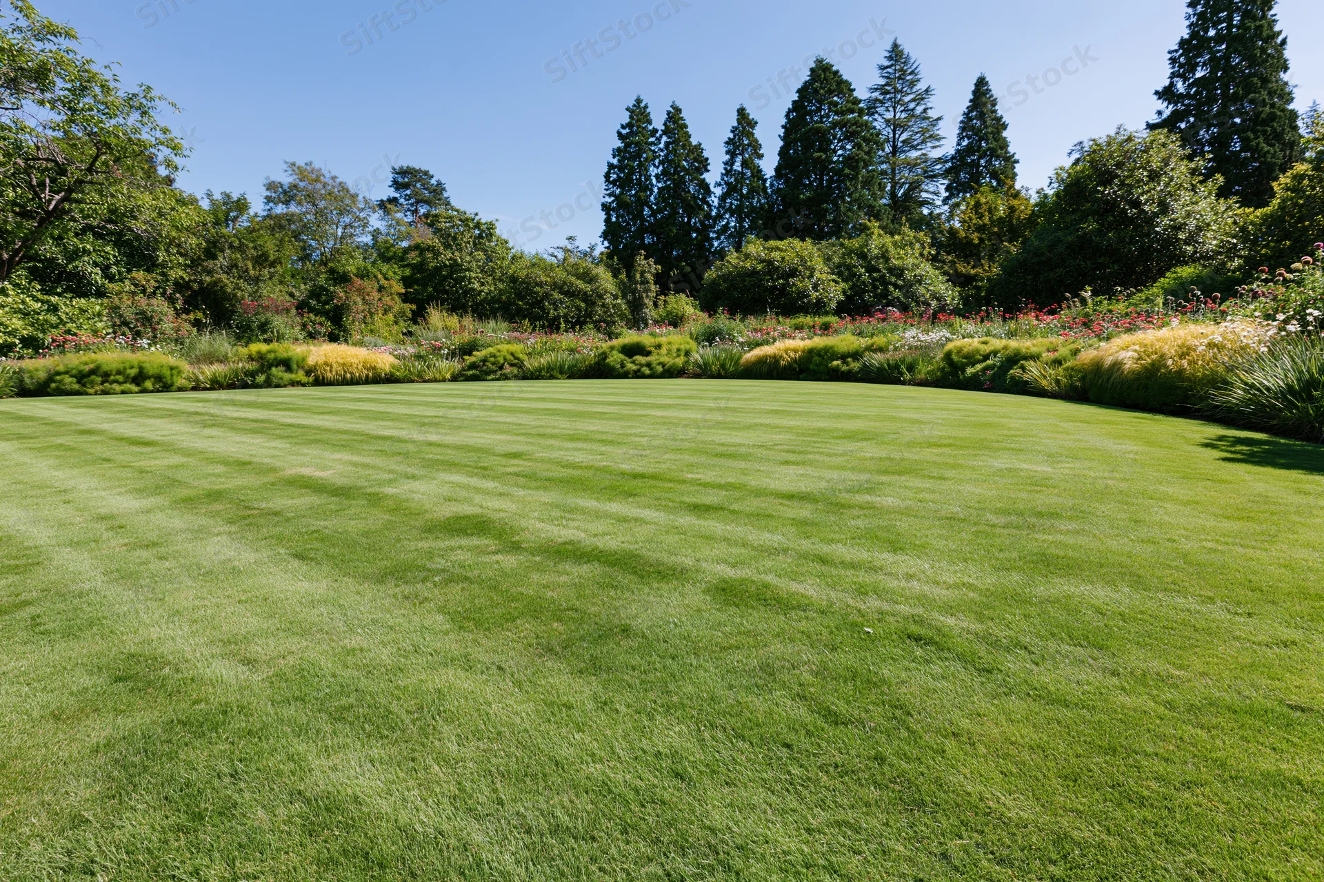 Expansive manicured lawn in a formal garden with striped grass and flowerbed borders under clear blue sky