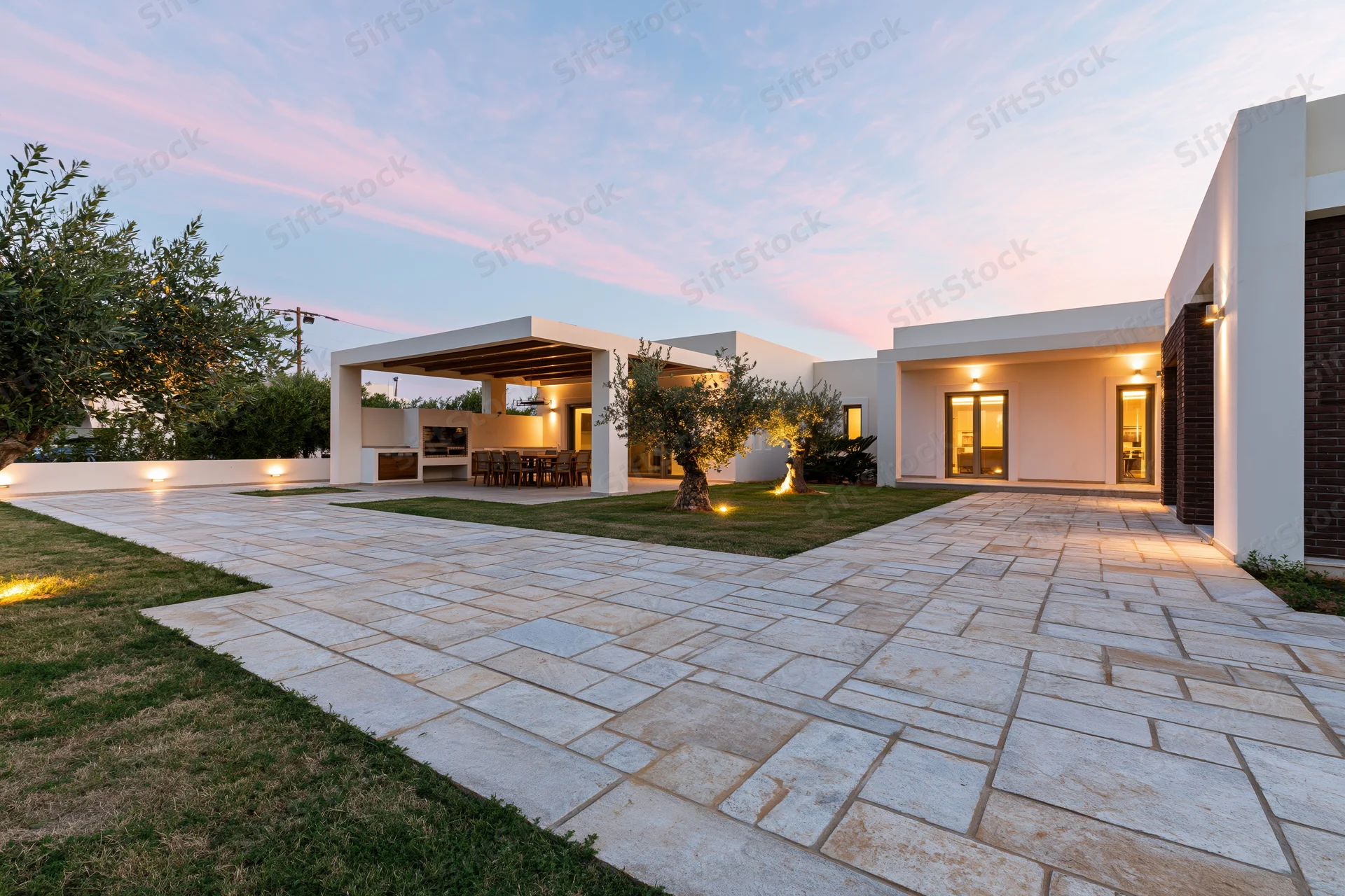 Modern villa exterior at dusk with stone pathway, patio, and olive trees in landscaped garden
