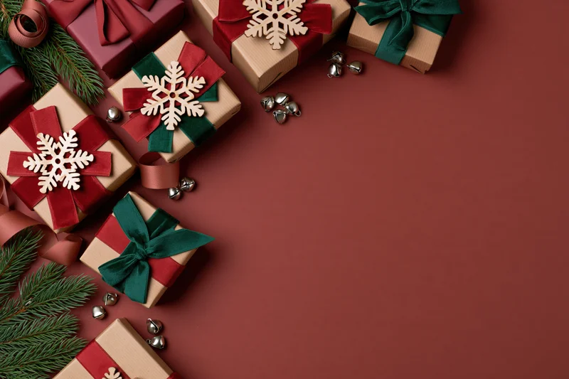 Christmas gift boxes wrapped in kraft paper with red and green ribbons, snowflake ornaments, pine branches, and jingle bells arranged in flat lay on burgundy background with copyspace