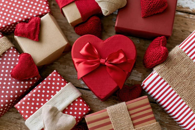 Heart-shaped gift box with red ribbon surrounded by wrapped presents on wooden surface