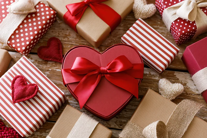 Heart-shaped gift box with red ribbon among assorted wrapped presents on wooden surface