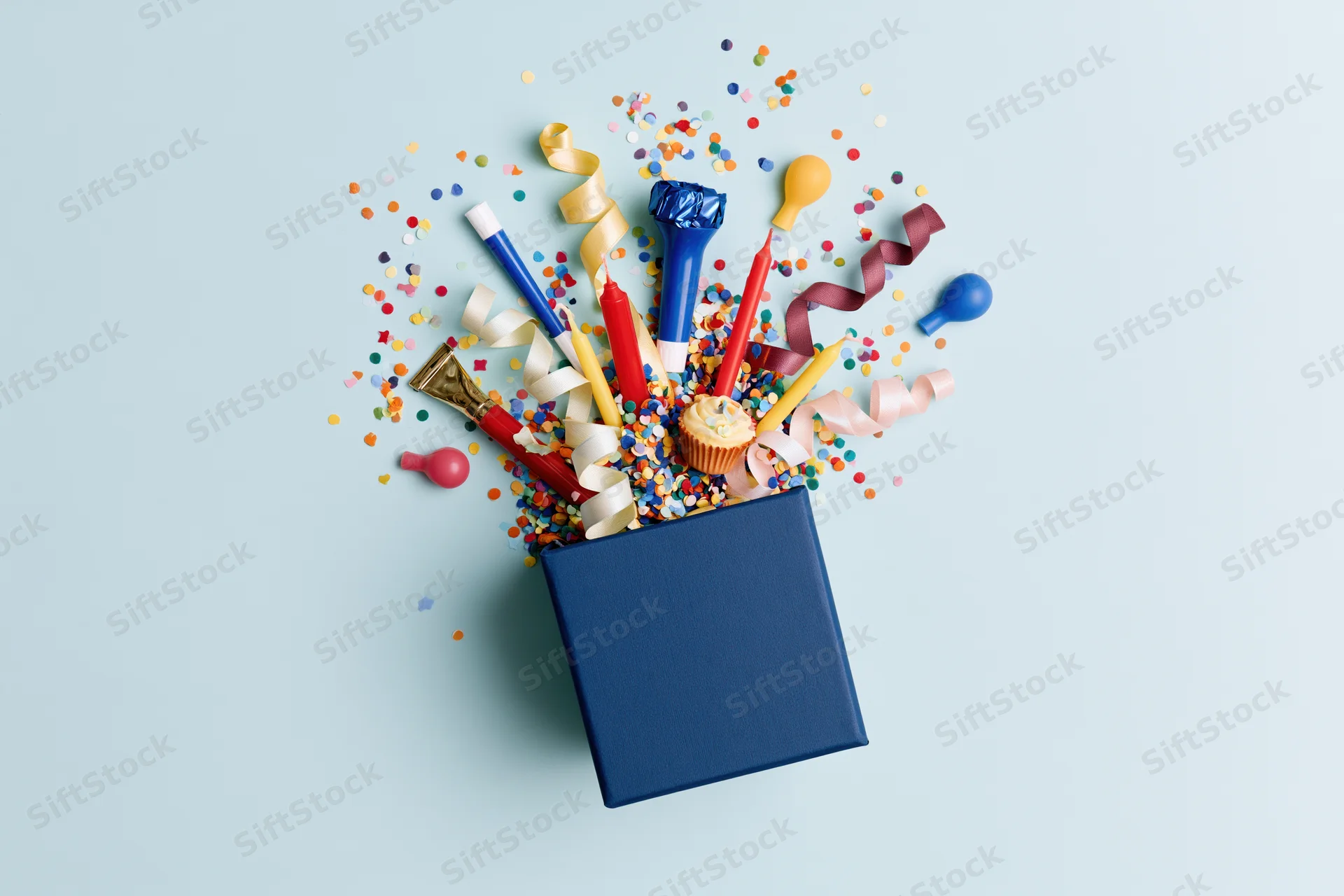 Dark blue gift box overflowing with birthday party items on light blue background