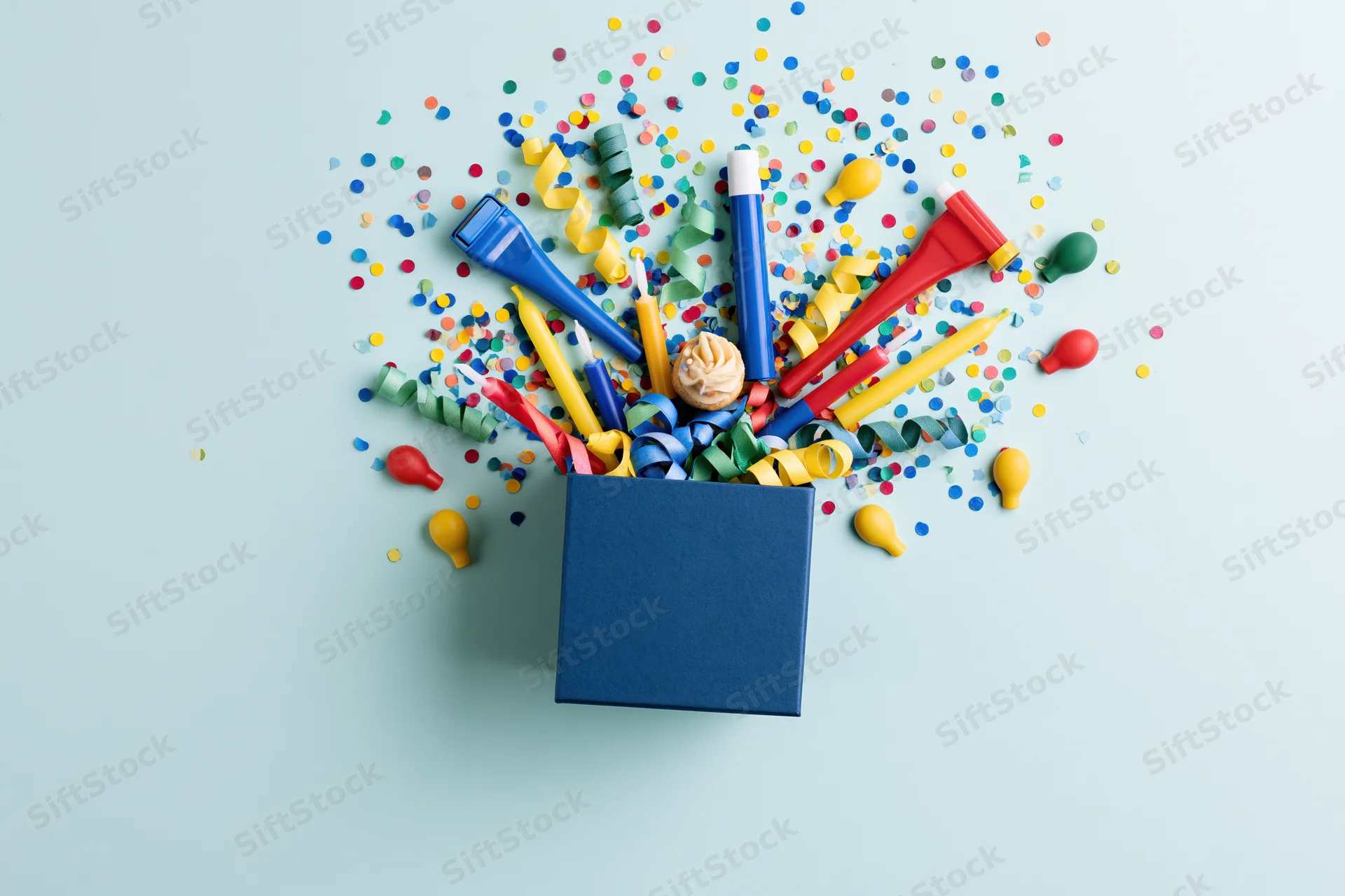 Dark blue gift box overflowing with birthday party items on light blue surface