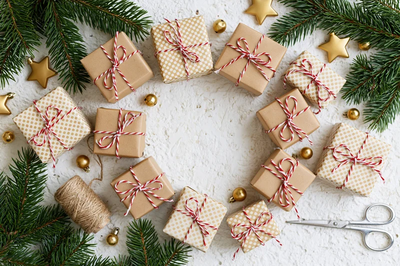Small christmas gift boxes wrapped in kraft and polka dot paper with red white twine bows arranged on white textured surface with pine branches and gold ornaments