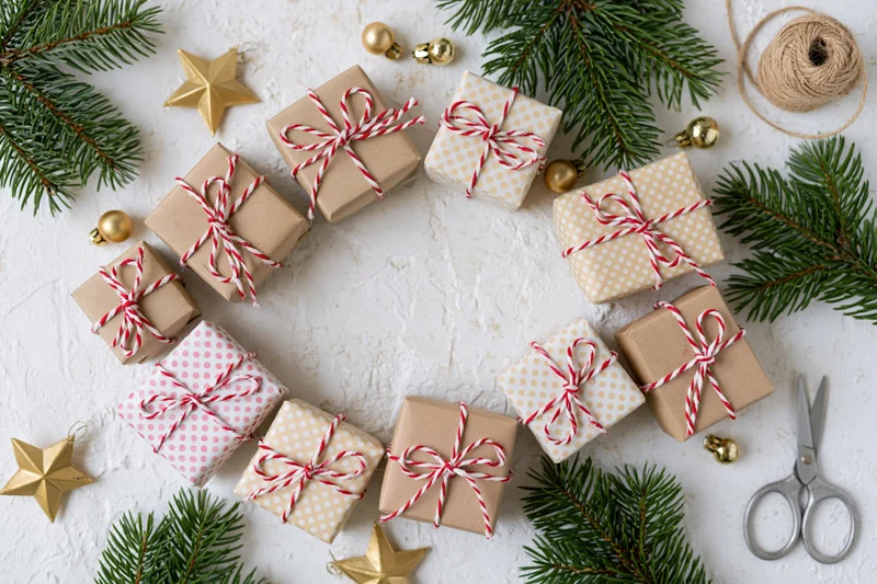 Small christmas gift boxes wrapped in kraft and patterned paper with red white ribbon arranged in circle on white surface with pine branches and ornaments