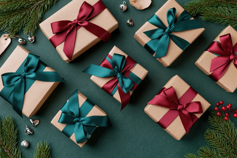 Gift boxes wrapped in kraft paper with teal and crimson ribbons arranged on dark green surface with pine sprigs and silver bells