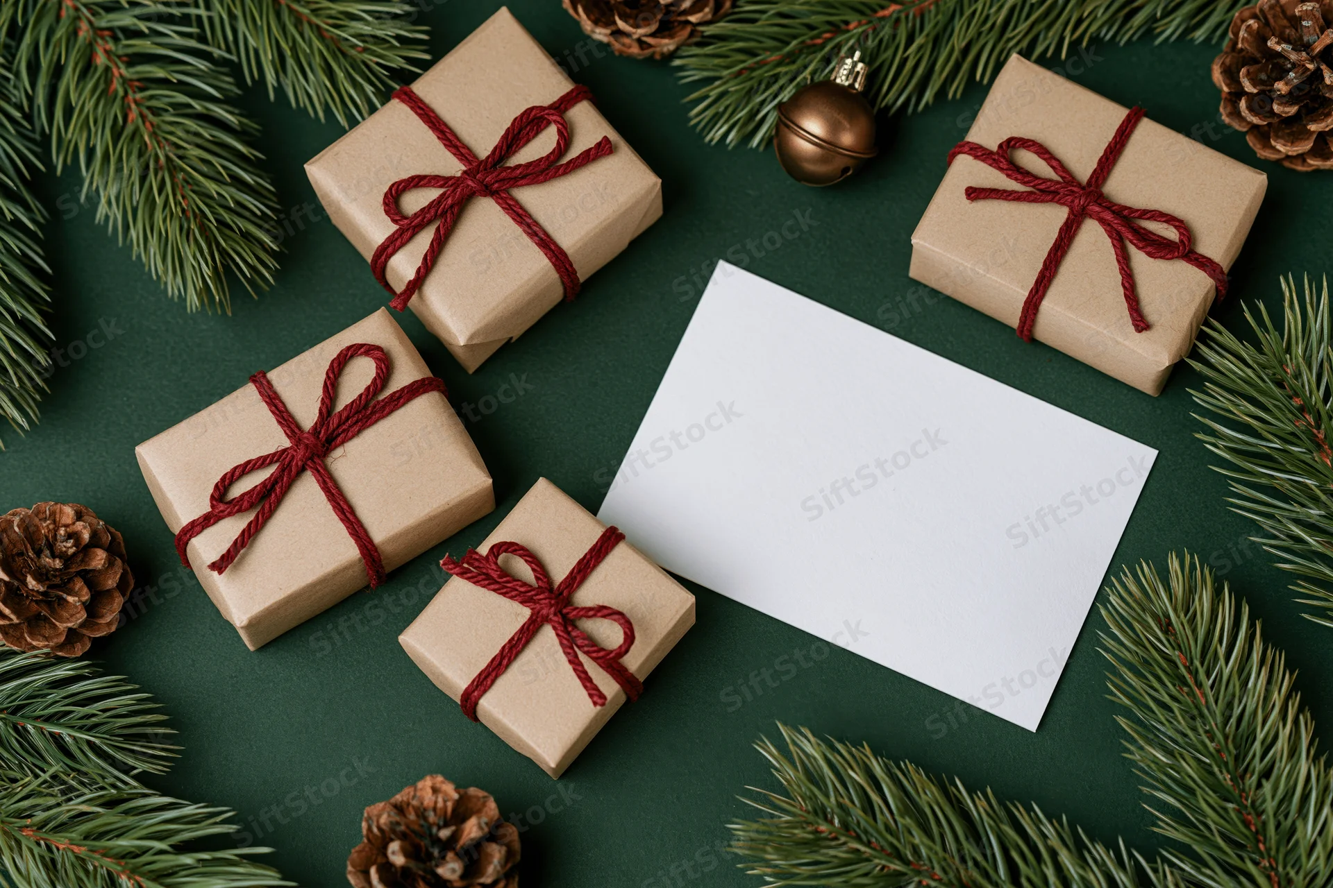 Four kraft paper gift boxes tied with red twine arranged with pine branches and pinecones on a dark green surface with a blank white card in the center