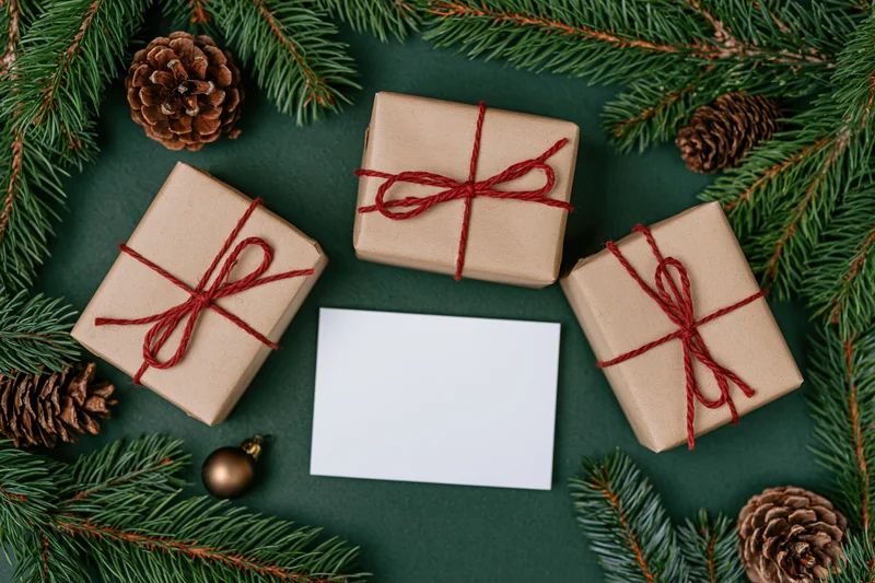 Three kraft paper gift boxes tied with red twine arranged with pine branches and pinecones around a blank card on dark green surface