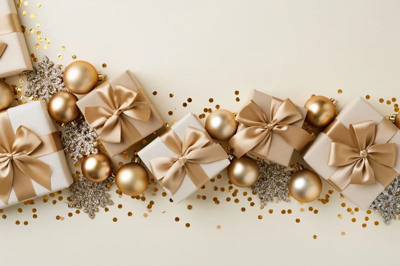 Wrapped gift boxes with gold satin bows and ornaments arranged in a flat lay on a light surface with confetti and snowflakes
