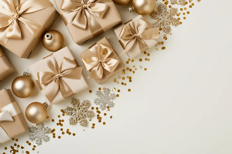Wrapped gift boxes with gold satin ribbon bows and holiday ornaments arranged in flat lay on white surface with copyspace