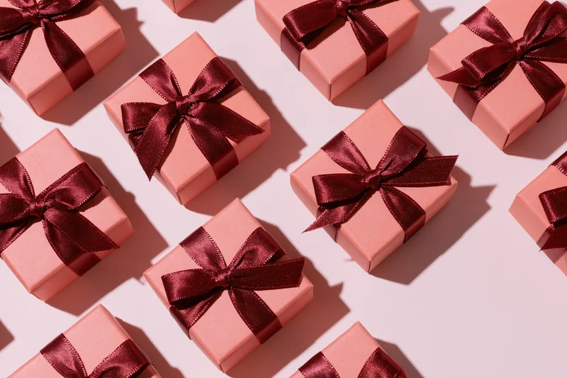 Coral-pink gift boxes with dark red satin bows arranged in a flat lay overhead pattern on a light pink surface