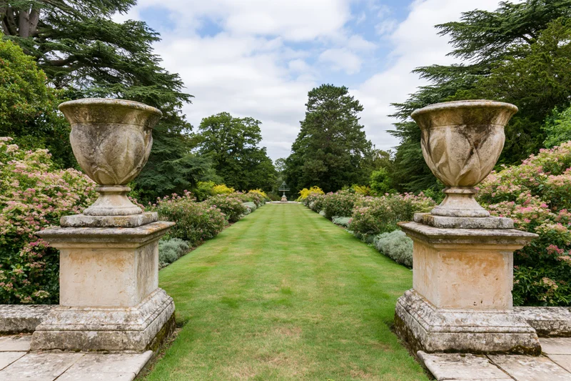Formal english country garden with stone urns on pedestals flanking a manicured lawn pathway leading to a distant fountain