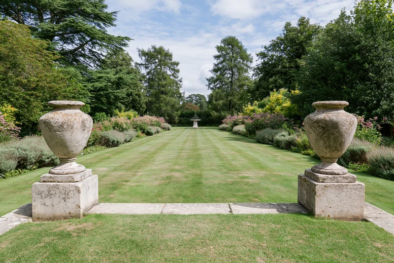 Formal english country garden with symmetrical lawn pathway flanked by stone urns on pedestals
