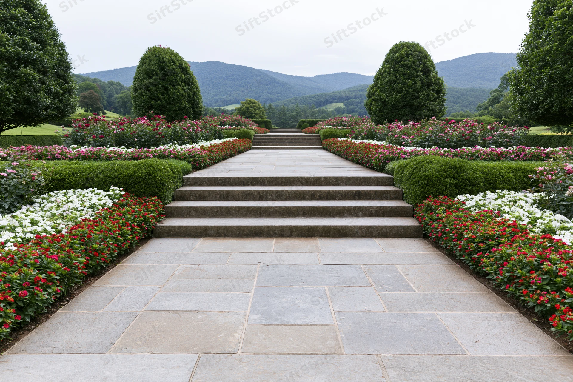 Wide stone steps leading through a formal garden with symmetrical flower beds and topiary hedges, framed by distant mountains