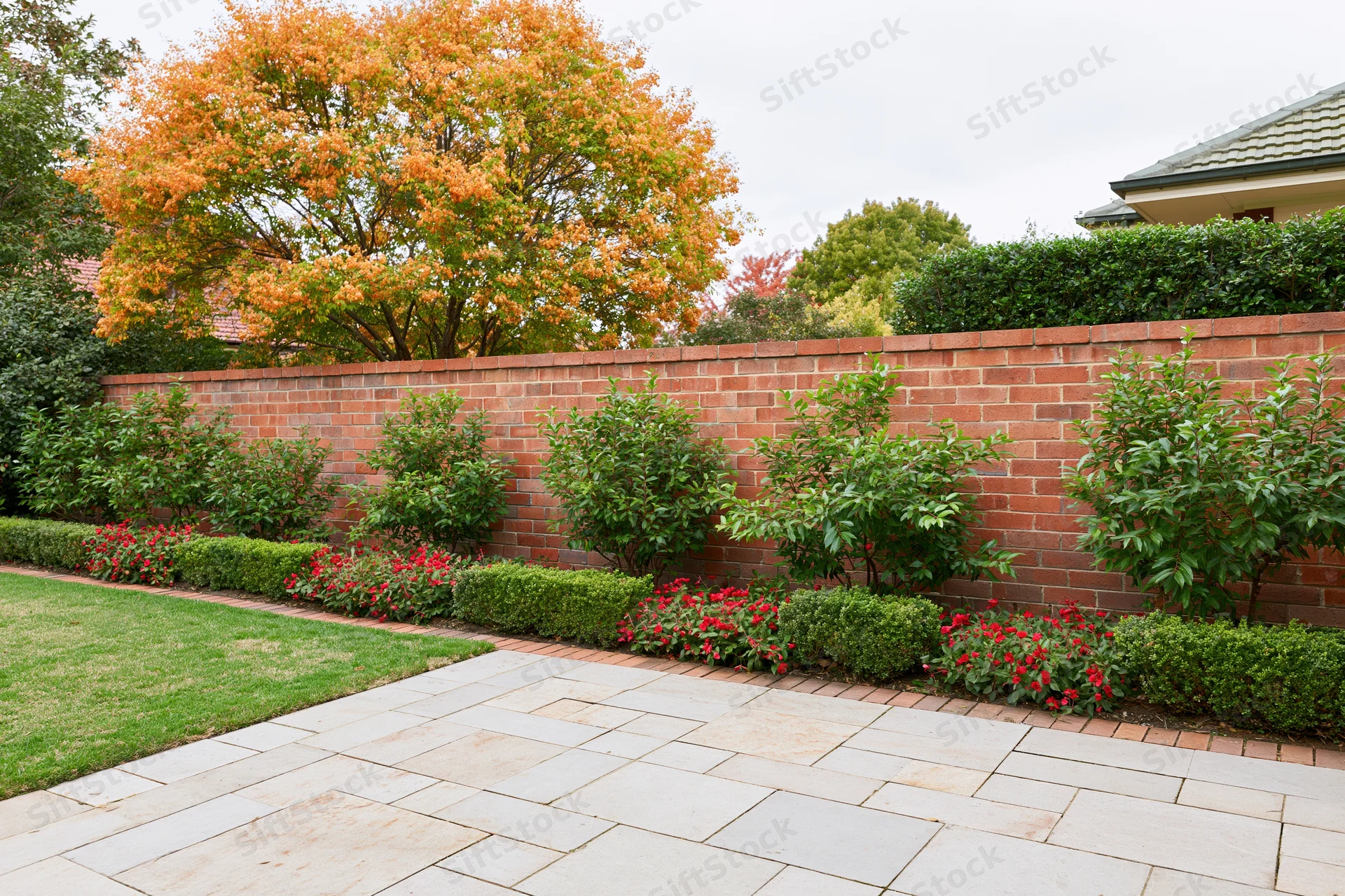 Formal garden with low red brick wall bordered by trimmed green shrubs and red flowers along stone patio and lawn