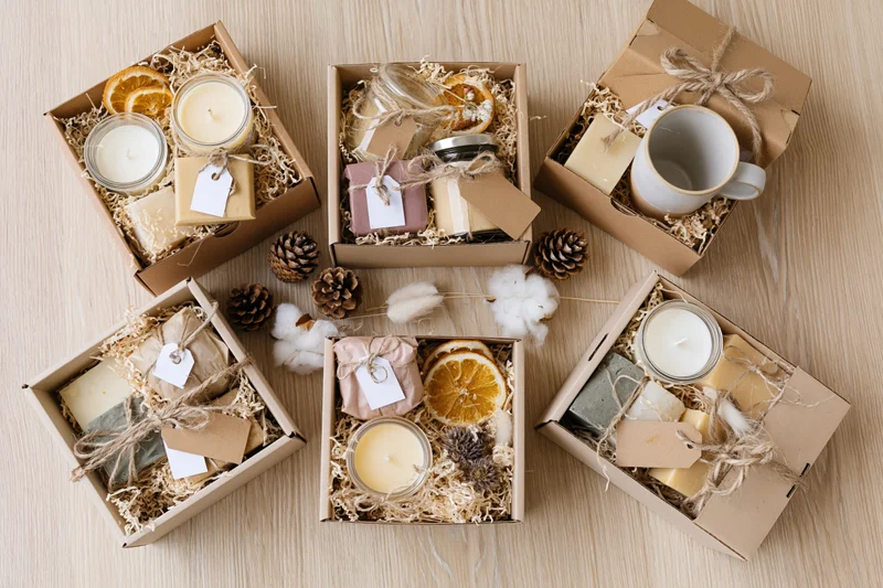 Open kraft gift boxes with candles, soaps, dried oranges and pinecones arranged on light wood surface