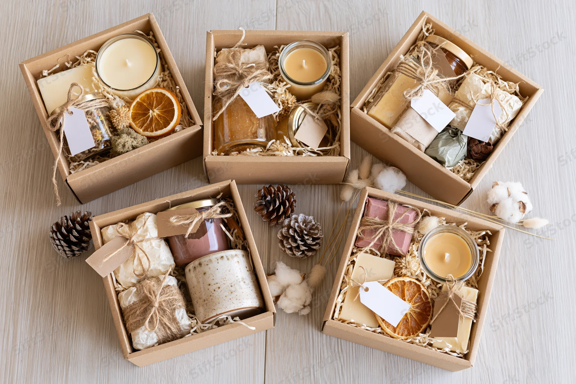 Five open kraft gift boxes filled with candles, soaps, and natural decor arranged on light wood surface