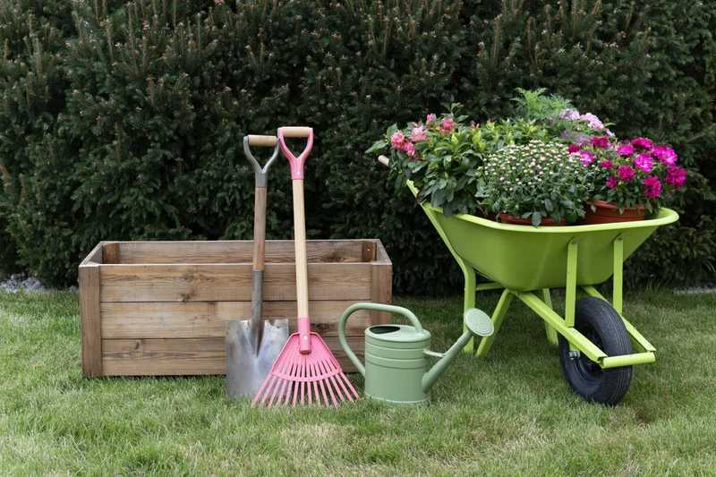 Garden tools and potted flowers arranged on a lawn beside a wooden planter box