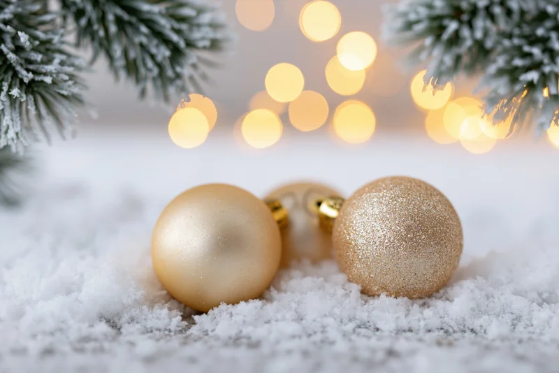 Gold christmas ornaments resting on snow with pine branches and bokeh lights in background