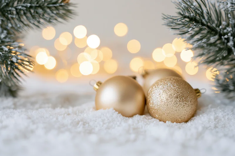 Gold christmas ornaments resting on snow with pine branches and blurred lights in background
